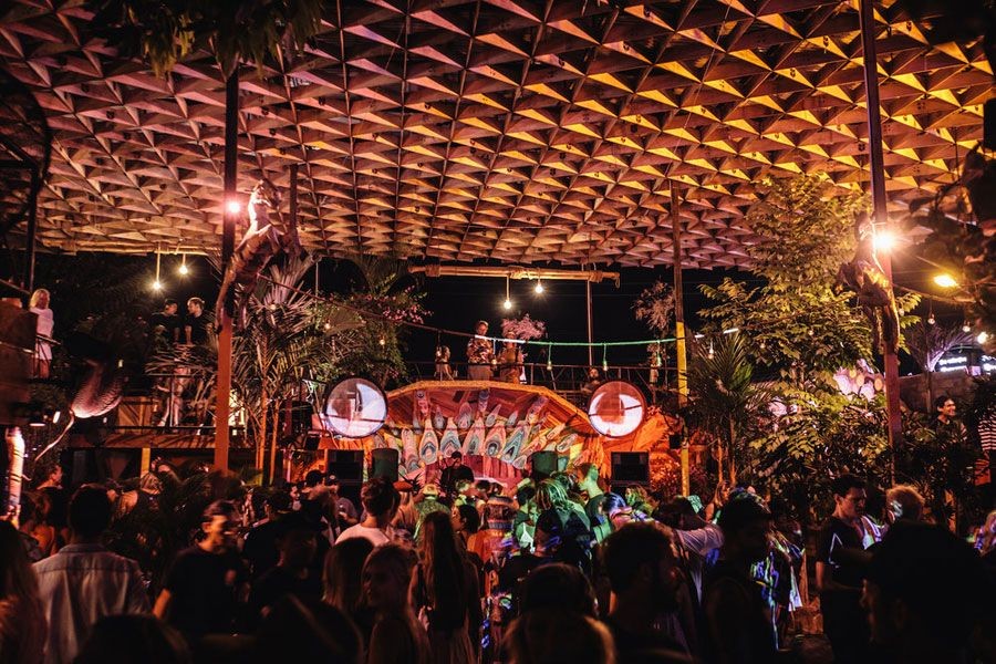 Crowded outdoor night party with people dancing under a geometric-patterned canopy and vibrant stage decorations.