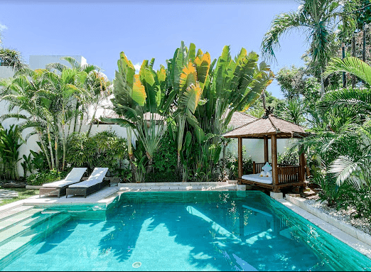 Tropical outdoor pool area with two lounge chairs, lush greenery, and a wooden gazebo.