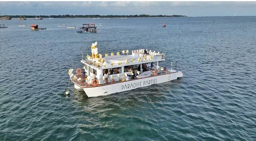 Party boat decorated with balloons, floating on a calm sea with several other boats in the background.