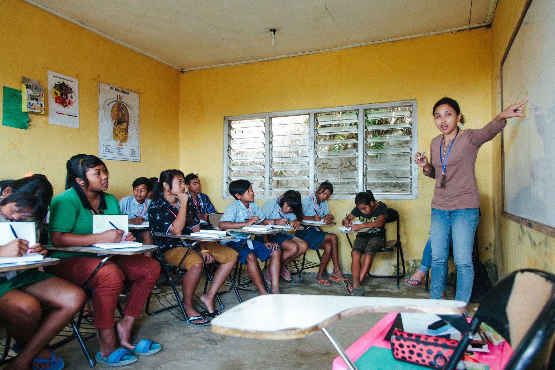 Teacher pointing to a whiteboard in a classroom while students sit at desks taking notes.